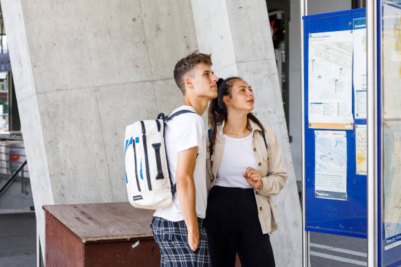 2 junge Menschen am Bahnhof vor Reiseinfowand 